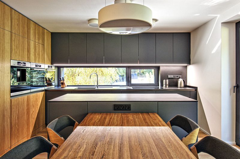 Panoramatické okno v kuchyni poskytuje výhled do přilehlé části zahrady a účinně osvětluje pracovní plochu. Také v kuchyni se opakuje motiv kombinace šedé barvy a dřeva