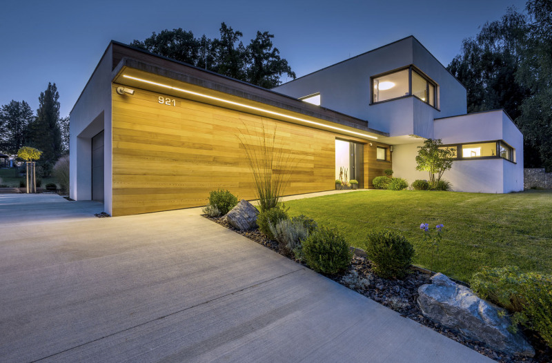 Garáž je orientována kolmo k hlavní obytné části domu. Podélně orientovaný obklad z olejovaného cedru a stříška s integrovaným LED osvětlením zvýrazňují horizontální hmotu a vedou příchozí k hlavním vchodovým dveřím