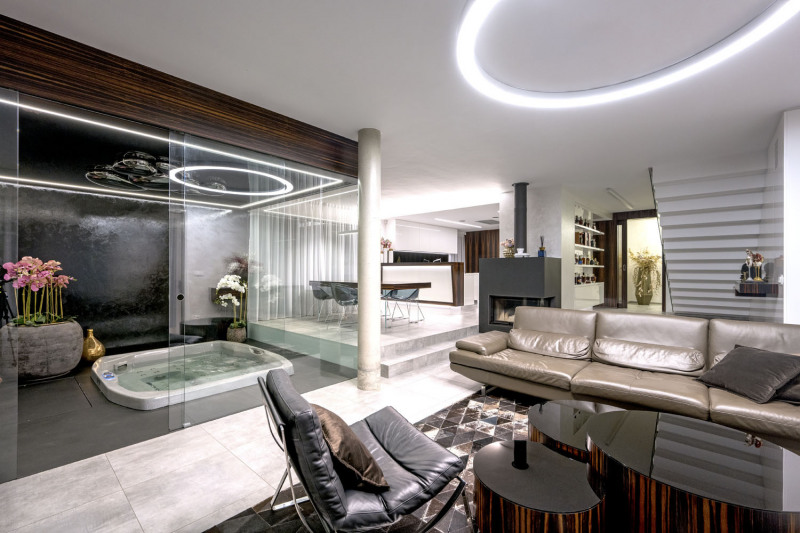 Společný obývací prostor tvoří otevřené zóny: kuchyň, jídelna, sezení, knihovna se schodištěm a wellness, oddělené skleněnou stěnou. Propojuje je krásná dlažba imitující travertin a vzácná makassarová dýha, která zdobí kuchyňskou linku, podhled vířivky, jídelní stůl a další solitéry