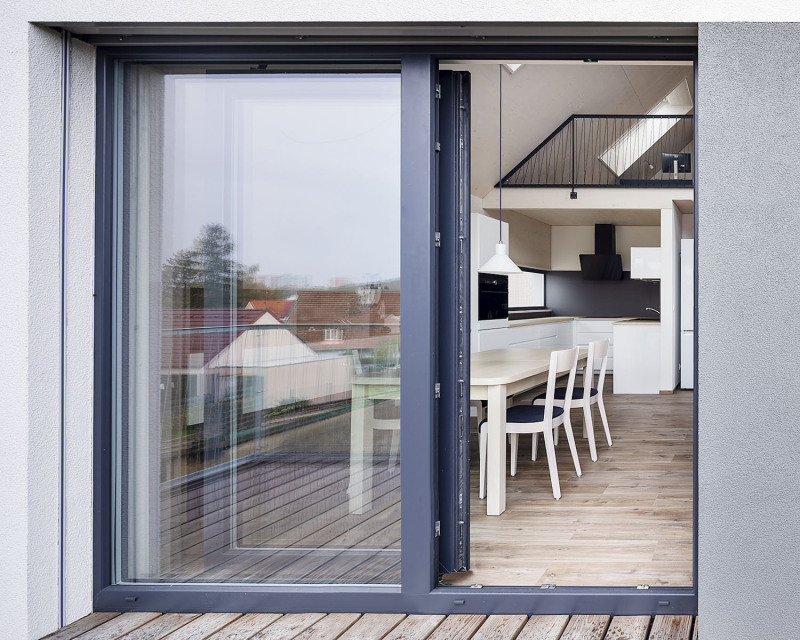 Průhled z terasy do jídelny spojené s kuchyní a obývacím pokojem. Celý prostor je otevřený do krovu, vzadu je vidět galerie s pracovnou osvětlenou také střešním oknem